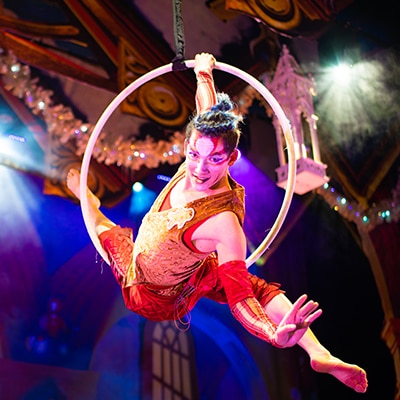 Acrobat performing an aerial hoop routine mid-air, wearing a vibrant red and gold costume with makeup