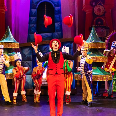 Performer in a red suit juggling hats on stage, surrounded by a colorful ensemble cast in circus-themed costumes during a vibrant theatrical performance.